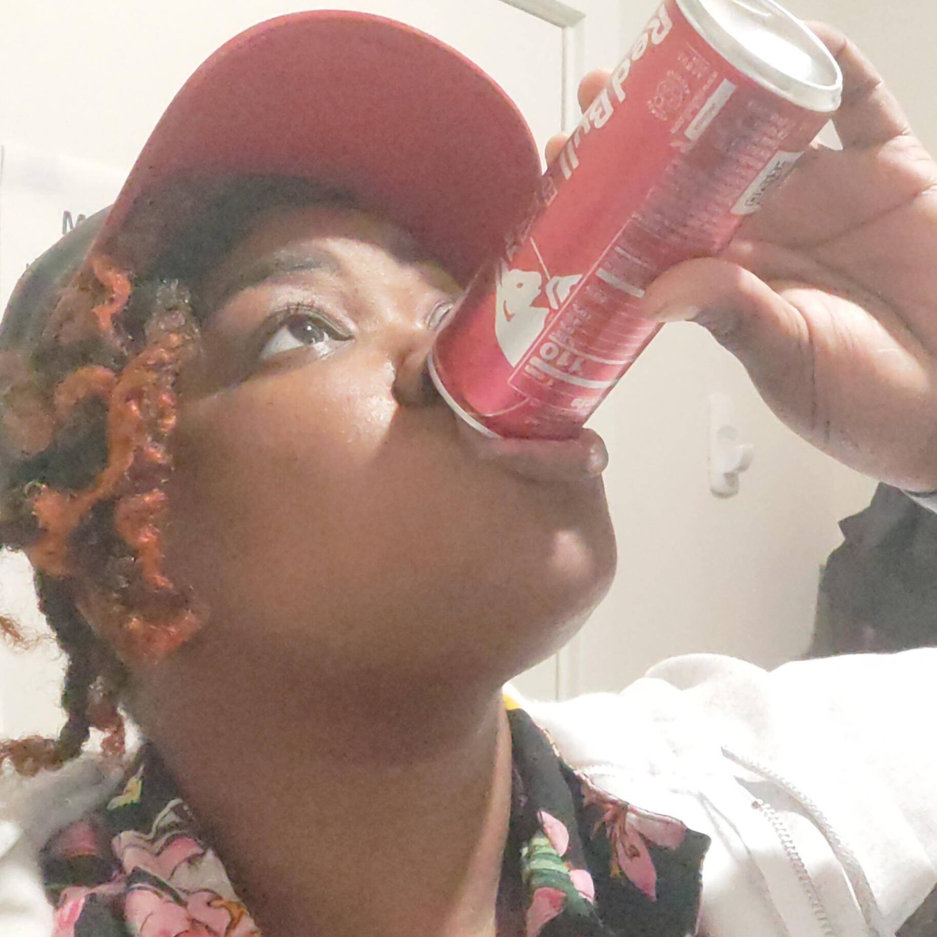 Dann, a collage aged Afro-Indigenous non-binary person w/dark brown skin and Black to Orange Ombre twisted hair under a baseball cap, is drinking a Redbull at a sharp angle with eye wide open.