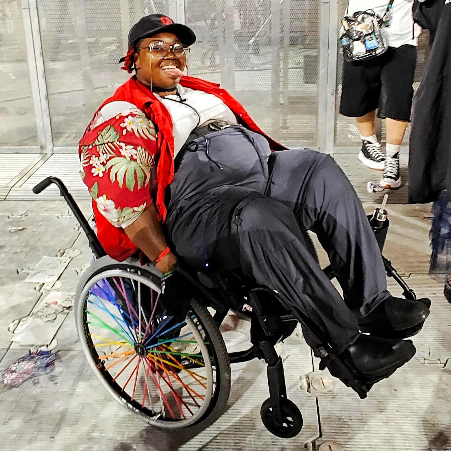 A photo of Dann, a dark skin afro indigenous person holding a wheelie in a wheelchair with rainbow spokes. They are wearing gray high-waisted pants, an open red pattern button up, a red vest, and a white shirt and black baseball cap. They are wearing glass
